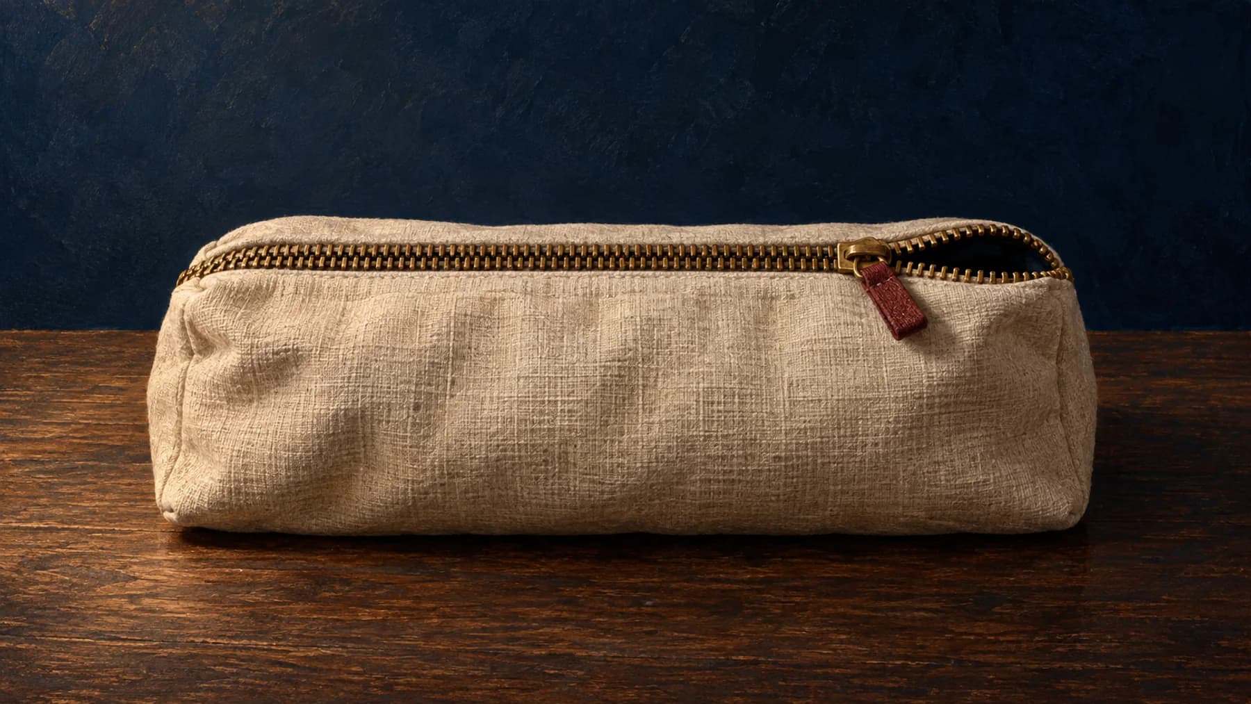 A cream linen pouch rests on walnut with its aged-brass zip almost closed, leaving a dark opening beside an oxblood pull-tab.