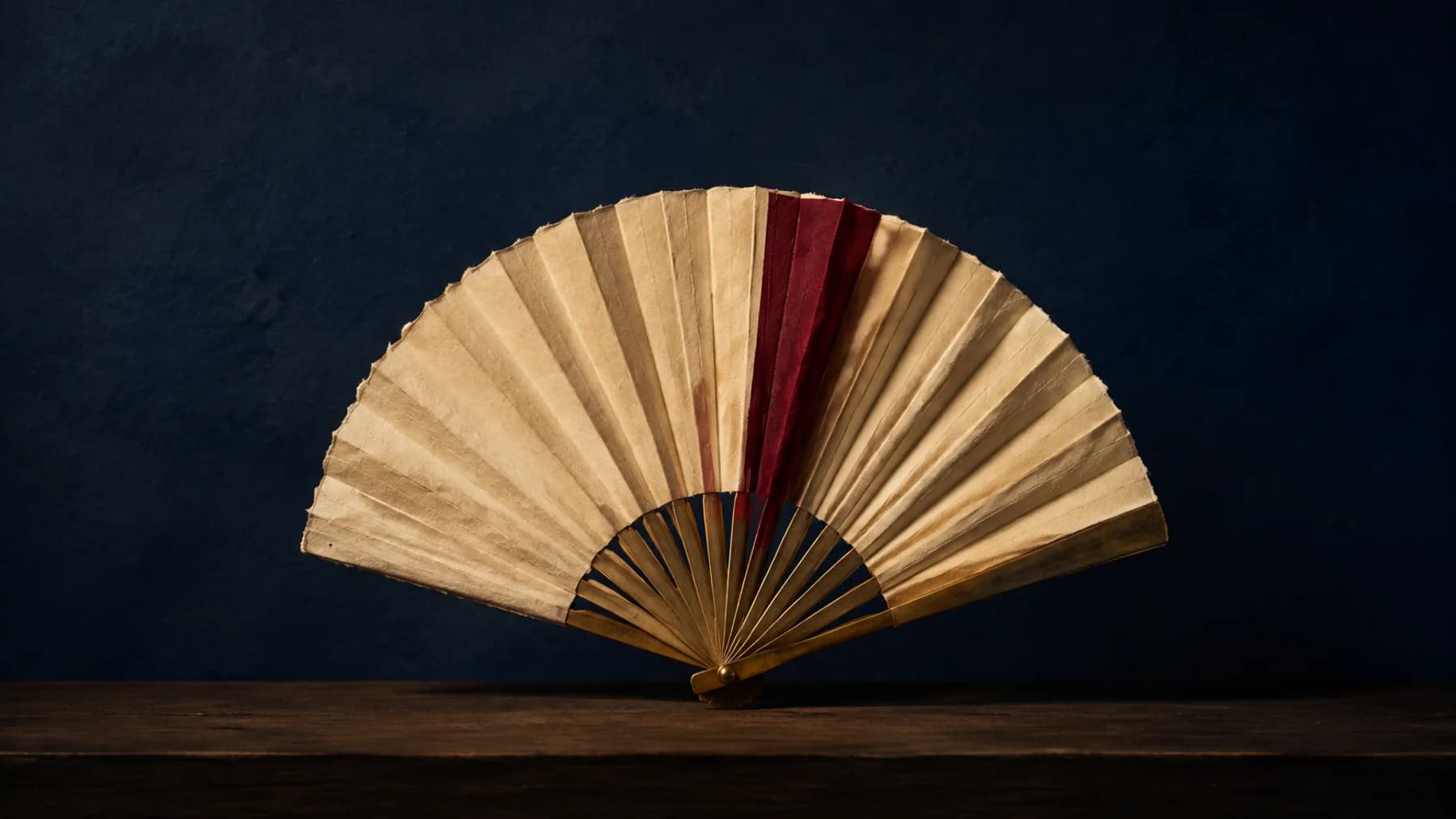 A cream handmade-paper fan with aged-brass ribs and one oxblood fold opens above a walnut shelf against midnight blue.
