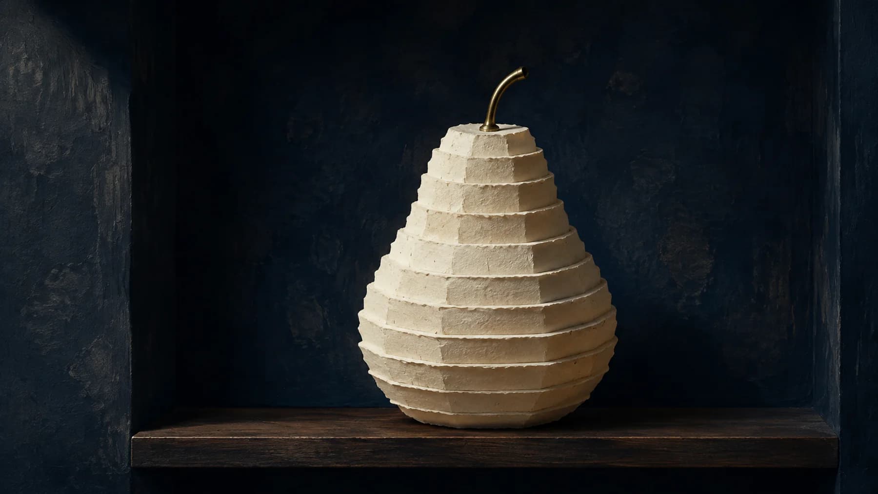 A pear built from stepped layers of cream handmade paper, with an aged-brass stem, rests on a walnut ledge against midnight blue.